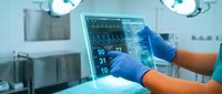 Medical professional in blue gloves operating patient monitoring system displaying vital signs in modern hospital operating room with surgical lights.