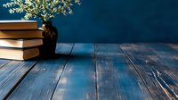 Stack of vintage books and white wildflowers in dark vase on rustic blue wooden table against deep blue wall background, creating cozy reading atmosphere.