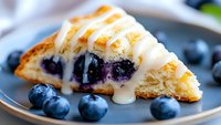 Fresh baked blueberry scone with vanilla glaze drizzle on gray plate, garnished with ripe whole blueberries, extreme close up showing moist texture.