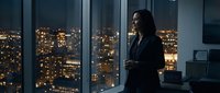 Woman in business attire standing by floor-to-ceiling windows overlooking illuminated cityscape at night, holding smartphone, contemplative pose in modern office setting.