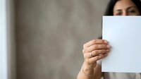 Woman with dark hair and medium skin tone holding blank white paper sheet in front of her face against neutral beige wall background for mockup and branding purposes.