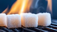 Sugar cubes on dark grill with bright orange flames and smoke in background, dramatic macro photography showing crystalline texture and heat contrast.