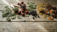 Assorted dried herbs and spices scattered on rustic wooden surface with copper measuring spoons for natural cooking and seasoning recipes.