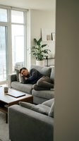 Young man relaxing on gray sofa in modern living room with natural light from large windows and green plant for home comfort and wellness lifestyle.