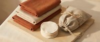 Eco-friendly zero waste bathroom set with folded linen towels, reusable cotton pads, and drawstring bag on wooden board in warm neutral tones.