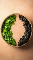 Fresh green tea leaves and dried black tea arranged in yin yang pattern inside round woven basket on brown background, showing tea processing transformation.