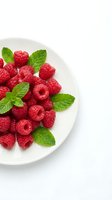 Fresh red raspberries with mint leaves arranged on white plate, isolated on white background. Suitable for food, nutrition, healthy eating, and recipe design projects.