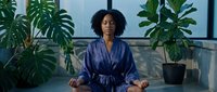 Woman with dark curly hair and medium skin tone meditating in lotus position wearing navy blue robe, surrounded by potted green plants in modern wellness studio space.