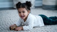 Happy mixed race toddler girl with curly hair in pigtails lying on textured rug, smiling joyfully while playing with wooden toy block in cozy home setting.