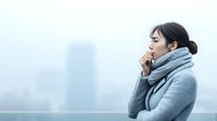 Young Asian woman in gray winter coat and scarf coughing outdoors against blurred cityscape background on cold foggy day, side view portrait showing illness symptoms.