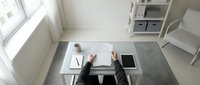 Professional workspace from above showing hands writing at white desk with tablet, coffee cup and notebook in bright modern office interior.