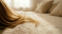 Flowing blonde hair cascades across soft beige bedding in warm natural light creating intimate morning atmosphere for beauty and wellness concepts.