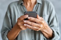Senior person with wrinkled hands holding smartphone, wearing light gray shirt against neutral background. Technology adoption by older generation.