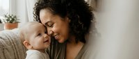 Happy mixed-race mother with curly hair bonding with her infant baby in cozy home setting, expressing love and maternal care during precious family moments.