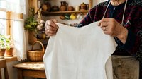Senior man with gray beard and weathered hands holding up clean white linen fabric in rustic kitchen with natural light, laundry and household care concept.