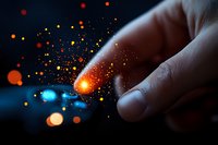 Magical glowing sparkles and light particles scatter from gentle finger touch against dark background, creating ethereal atmosphere with orange and blue bokeh effects.