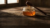 Golden honey dripping from wooden dipper into glass jar with twine bow on rustic wooden table in warm sunlight for natural organic food packaging.