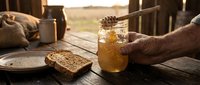 Man's hand holding jar of honey with bread on rustic wooden table outdoors, golden sunlight, countryside setting for food and agriculture themes.
