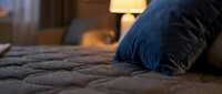 Cozy bedroom interior with navy blue pillow on quilted bedding, warm lamp lighting in background, comfortable home decor for relaxation and sleep design projects.