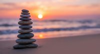 Balanced stone stack on beach at sunset with warm golden light reflecting on ocean waves creating peaceful zen meditation atmosphere for wellness.