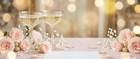Elegant champagne glasses with pink roses and baby's breath flowers on white table with golden bokeh lights background for wedding celebration.