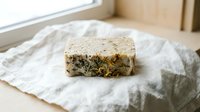 Handmade soap bar with dried herbs and natural ingredients on white linen cloth, organic skincare product for wellness and beauty routines.