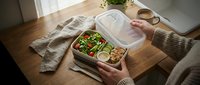 Overhead view of person in beige sweater holding open compartmentalized meal prep container with fresh salad, vegetables and protein portions on wooden table with napkin and accessories.