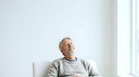 Mature man with gray hair and beard, Caucasian ethnicity, wearing casual gray sweater, sitting alone by bright window in contemplative pose, suitable for lifestyle, wellness, and editorial use.