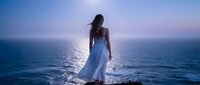 Young woman in flowing white dress standing on ocean shore at sunset with dramatic sky and shimmering water reflections creating peaceful meditation moment.