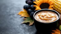Homemade pumpkin puree in black bowl with autumn decorations, corn, grapes and sunflower on dark background. Fall seasonal food photography.