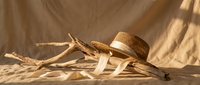Woven straw hat with ribbon and driftwood branches on sandy beach surface with warm sunlight and shadows creating summer vacation atmosphere.