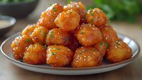 Crispy fried potato balls glazed in sweet and spicy orange sauce, garnished with fresh chopped herbs on gray ceramic plate, close up food photography.