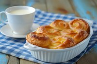 Fresh baked sweet bread pudding in white ceramic baking dish served with cup of hot milk on blue checkered napkin on rustic wooden table.
