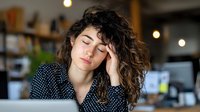 Young woman with curly hair feeling tired at workplace, resting head on hand while working on laptop in cozy cafe environment.