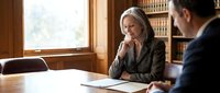 Mature woman with gray hair in professional business attire conducting formal meeting or interview with male colleague in traditional office setting with bookshelves.