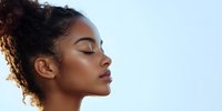Young African American woman with natural curly hair in profile view against light background, eyes closed, showing glowing skin and serene expression.