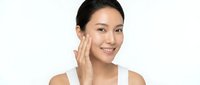 Young Asian woman applying skincare cream to face with gentle touch, smiling confidently on white background for beauty and wellness campaigns.