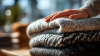 Hand resting on stack of folded cozy woolen sweaters in warm sunlight, showcasing soft textures and natural fibers for winter fashion.
