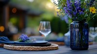 Romantic outdoor table setting with blue glass vase filled with wildflowers, woven placemat, wine glass and blurred garden background in warm evening light.