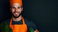 Smiling Caucasian male chef in orange apron and hat holding fresh green vegetables against dark background. Professional food service and healthy cooking concept.