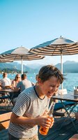 Young Caucasian man enjoying cold beer at lakeside restaurant terrace with striped umbrellas and mountain views during summer vacation.