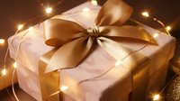 Elegant gift box wrapped in soft pink paper with luxurious golden satin ribbon bow, surrounded by warm glowing fairy lights creating festive holiday atmosphere.