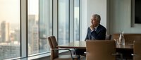 Middle-aged man with gray hair and light skin tone, wearing business attire, sitting at desk in modern high-rise office with city skyline view, working on laptop during daytime.