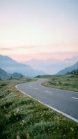 Winding asphalt road curves through alpine valley with misty mountains at dawn, surrounded by green meadow grass and wildflowers for travel and landscape design.