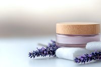 Pink cosmetic cream jar with wooden lid alongside fresh lavender flowers and white cotton pads on light background, spa and skincare concept.