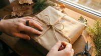 Person wrapping kraft paper gift box with twine bow, hands tying present with natural decorations and scissors on wooden table.