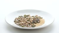 Assorted raw seeds and grains arranged on white plate including sunflower seeds, pumpkin seeds, chia seeds, and quinoa for healthy nutrition and cooking.