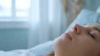 Young woman lying peacefully in bed with eyes closed, relaxing in bright morning sunlight streaming through window for wellness and sleep concepts.