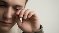 Young Caucasian man wiping tears from his eye, expressing deep sadness and emotional pain, close-up portrait on neutral background.