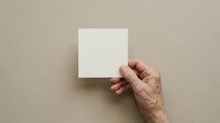 Mature adult hand holding blank white square card against neutral beige wall for mockup, branding, or presentation purposes.